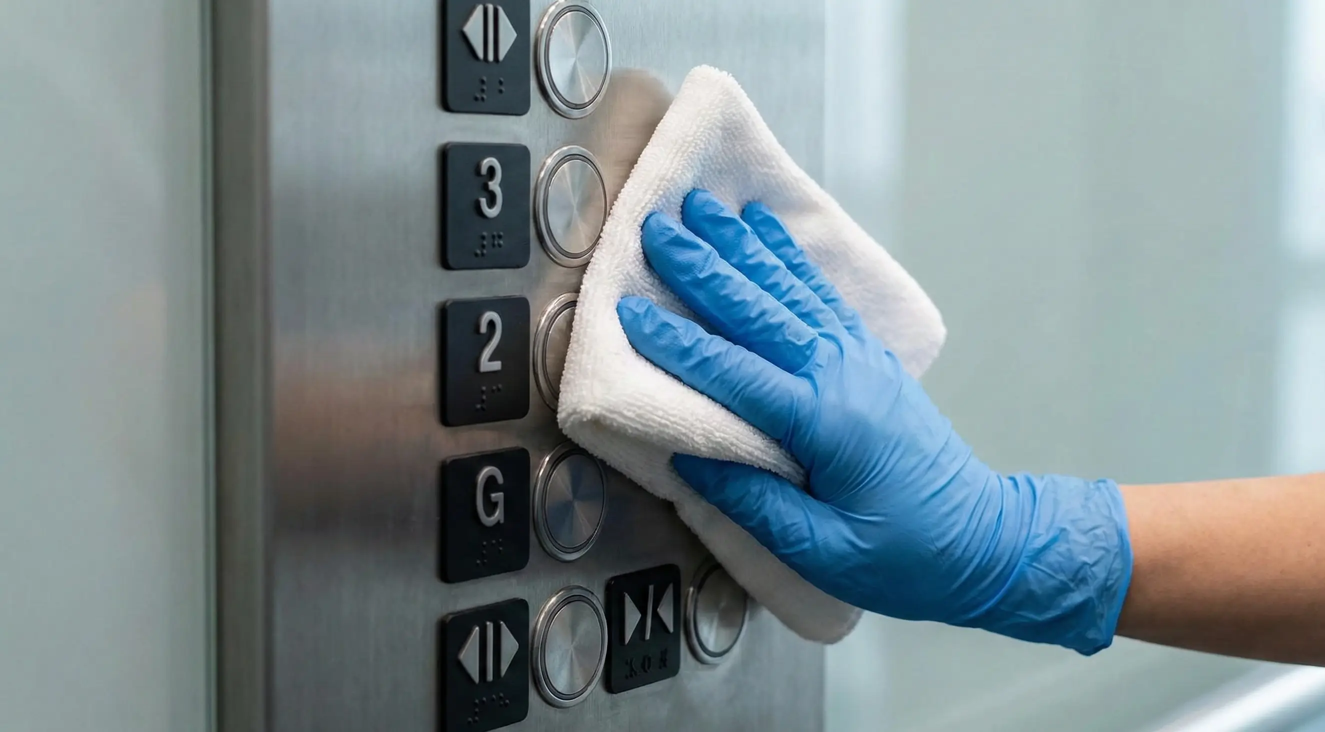 Close-up of a professional cleaner's gloved hand disinfecting high-touch elevator buttons in a busy office building using a microfiber cloth to prevent cross-contamination.